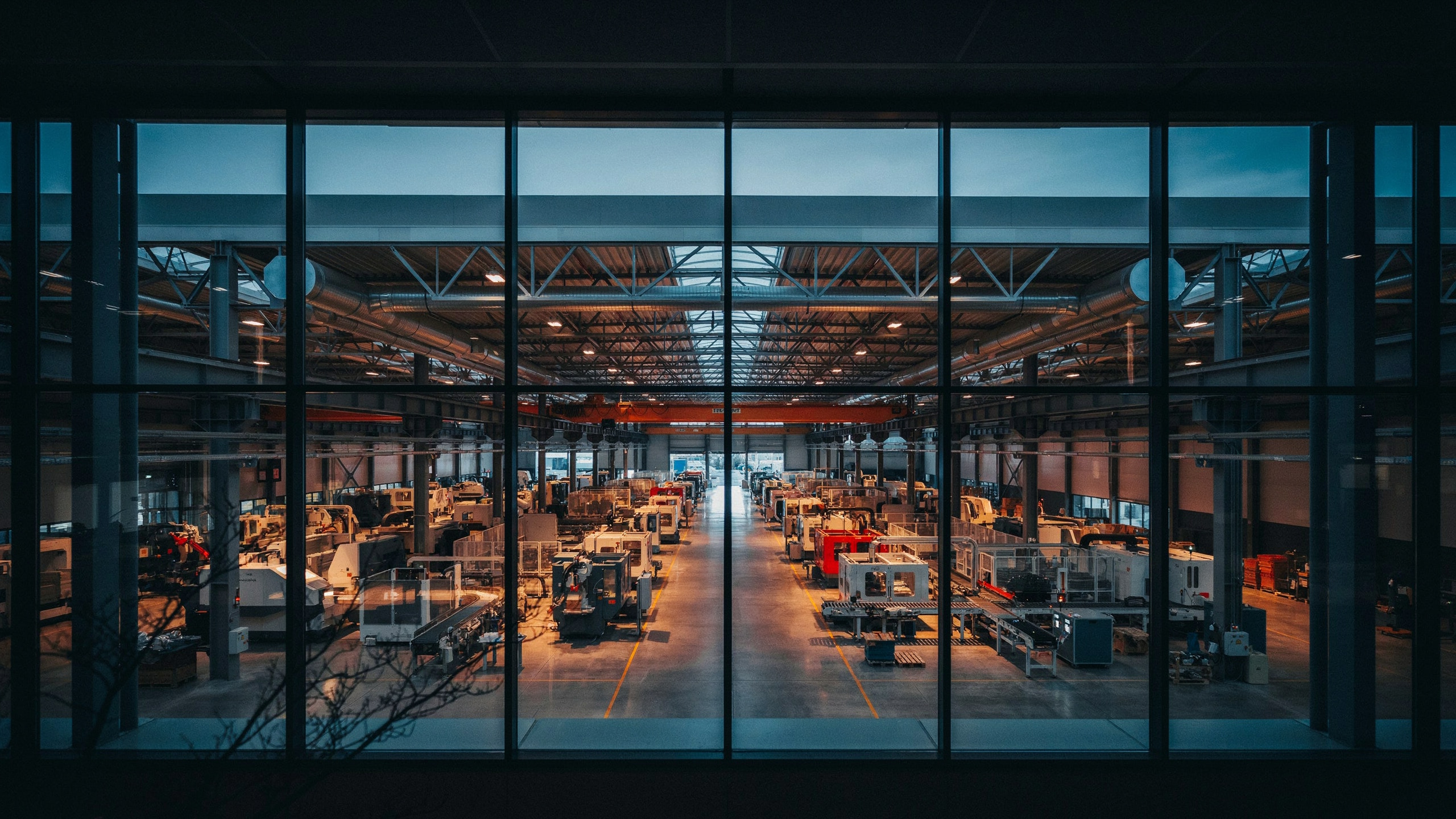 Large industrial factory interior at dusk seen through glass, warm amber light illuminating manufacturing equipment