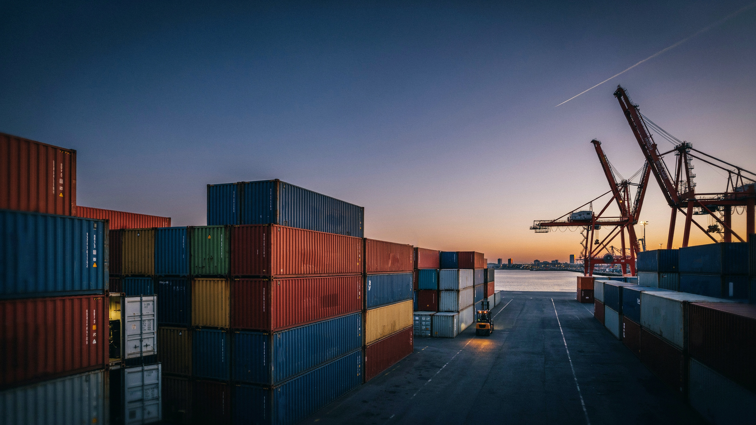 Modern European port terminal at dusk, shipping containers and cranes silhouetted against amber sky